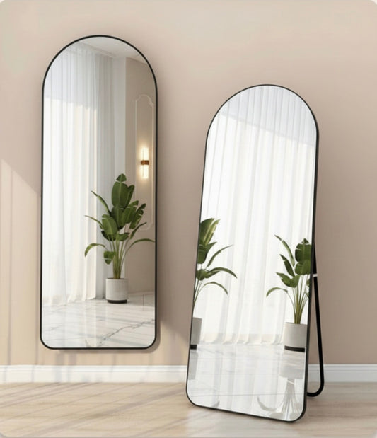 Two arched full-length floor mirrors with thin black frames, one standing against the wall and one leaning on a stand, showing a reflection of a potted plant and white curtains in a bright room.