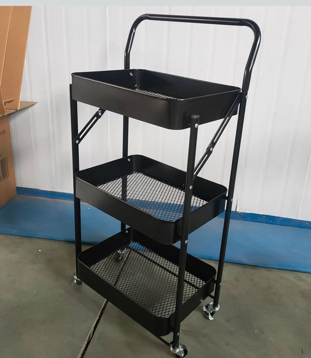 A three-tier black metal rolling cart with mesh shelves and a top handle, photographed indoors against a white paneled wall and blue baseboard.