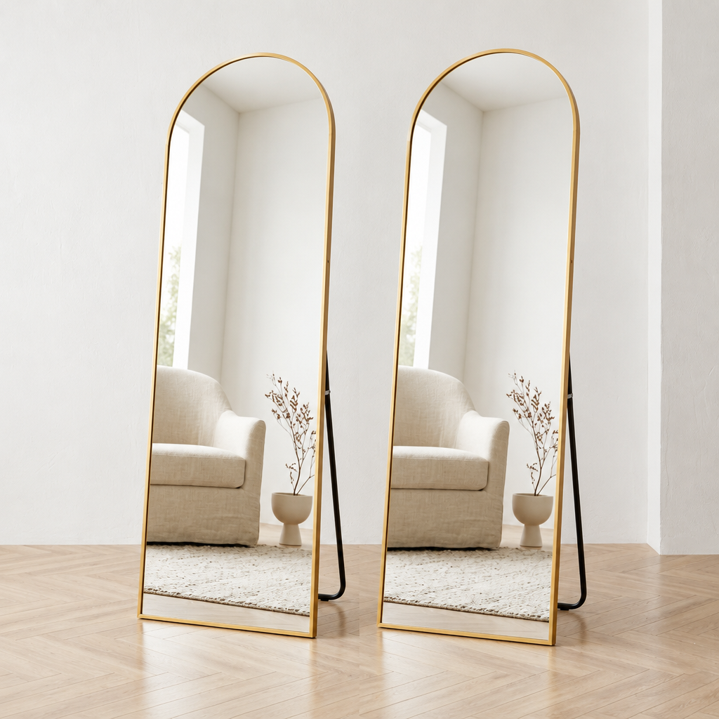 A tall arched full-length floor mirror with a slim gold metal frame and a black support leg, standing on a light wood floor against a white wall.