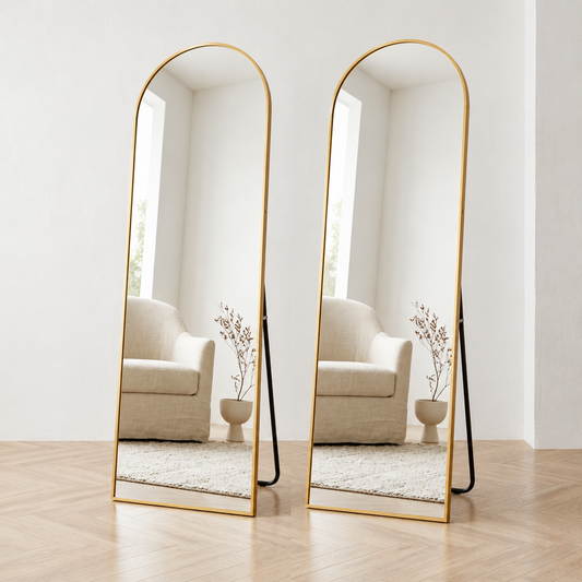 A tall arched full-length floor mirror with a slim gold metal frame and a black support leg, standing on a light wood floor against a white wall.