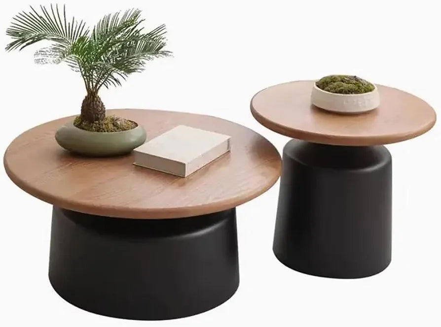 Two modern low coffee tables with round light wood tops and matte black cylindrical bases, shown on a white background with decorative props including a potted plant, a book, and a small bowl.