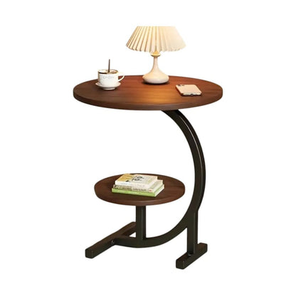 A modern round wooden side table with a dark curved metal frame and a lower shelf, styled with a table lamp, a cup of coffee, and books on a plain white background.