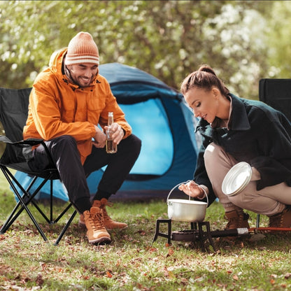 Two people camping outdoors, one holding a beer and the other using a portable stove.