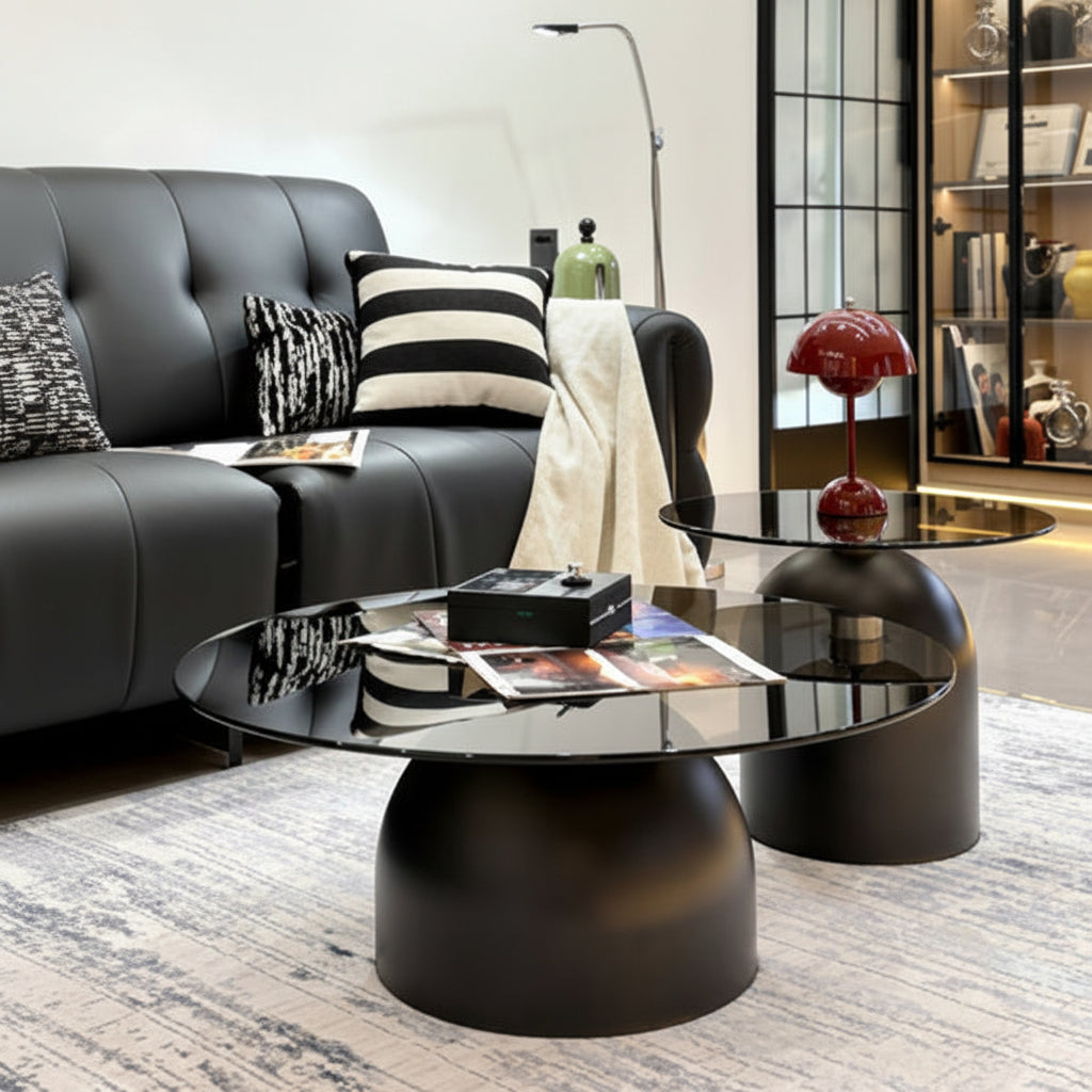 Modern round coffee table with a glossy black glass top and a matte black dome-shaped base, shown in a styled living room next to a black leather sofa.
