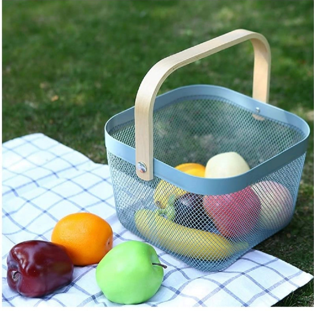 A gray mesh picnic basket with a light wood handle sits on a blue-and-white checkered blanket on grass, with fruit placed around it.