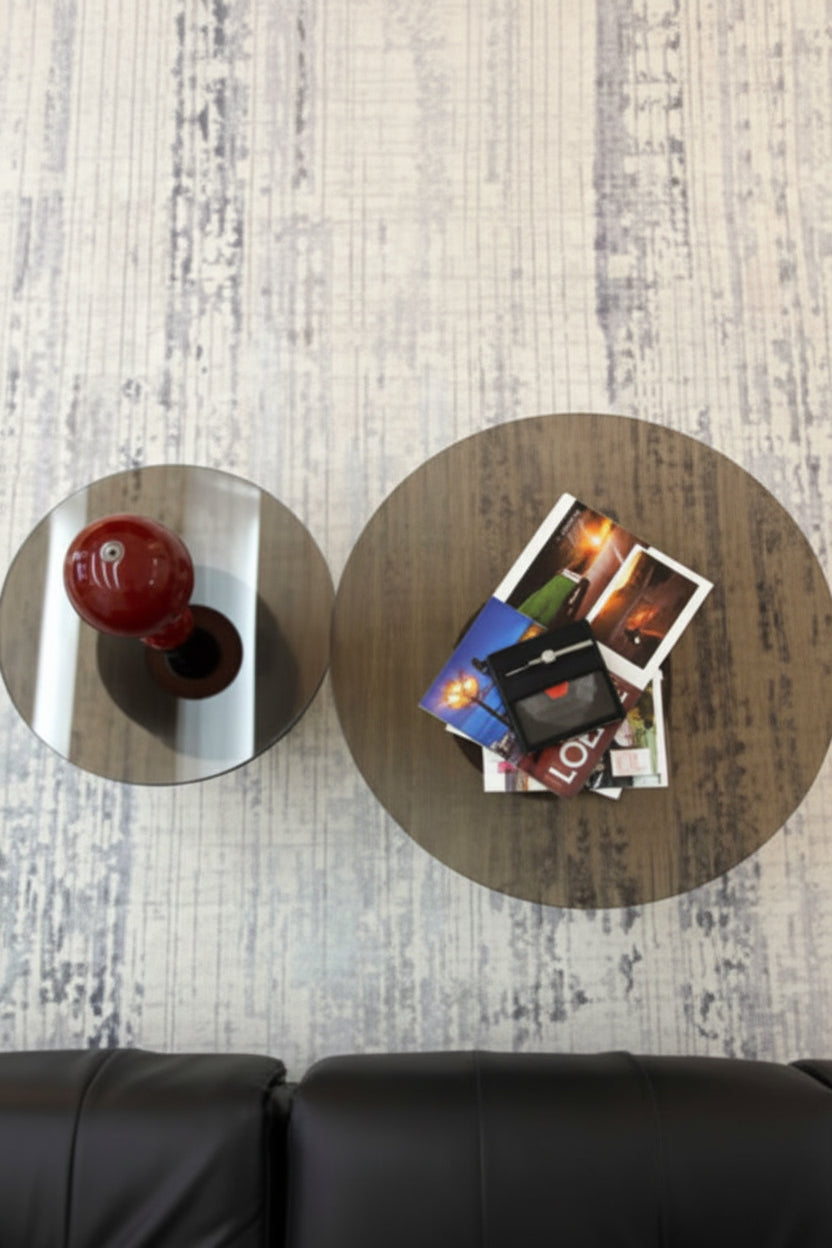 Two round coffee tables on a light wood floor, with a black leather sofa visible at the bottom. One table is mirrored and shows a red vase, while the other has a wood grain finish and holds a stack of Polaroid photos.
