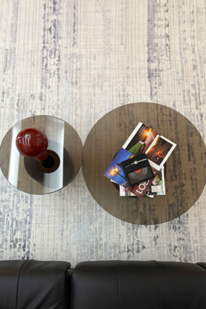 Two round coffee tables on a light wood floor, with a black leather sofa visible at the bottom. One table is mirrored and shows a red vase, while the other has a wood grain finish and holds a stack of Polaroid photos.